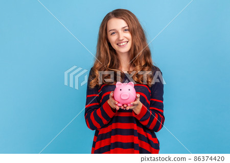 Beautiful woman wearing striped casual style sweater, holding pink piggy bank, looking at camera with charming smile, best variant for saving money. Indoor studio shot isolated on blue background. Beautiful woman wearing striped casual style sweater, holding pink piggy bank, looking at camera with charming smile, best variant for saving money. Indoor studio shot isolated on blue background. 86374420