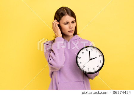 Portrait of confused woman holding in hands big wallclock, has no time , worried about deadline, looking away, wearing purple hoodie. Indoor studio shot isolated on yellow background. 86374434