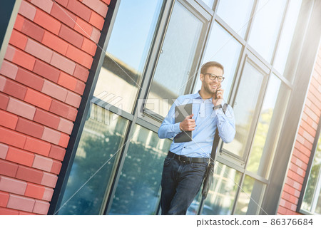 Young happy caucasian businessman wearing blue shirt and eyeglasses holding his laptop, talking by phone and smiling while walking outdoors Young happy caucasian businessman wearing blue shirt and eyeglasses holding his laptop, talking by phone and smiling while walking outdoors 86376684