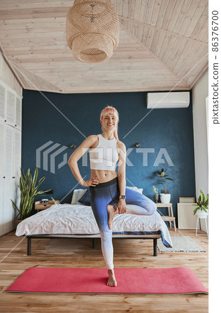 Smiling woman in sportswear does vrksasana holding leg in spacious room Smiling woman in sportswear does vrksasana holding leg in spacious room 86376700