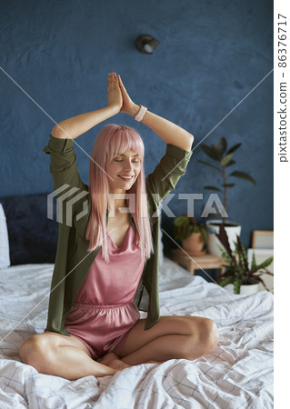 Graceful woman with colorful hair meditates holding hands in namaste above head on bed Graceful woman with colorful hair meditates holding hands in namaste above head on bed 86376717