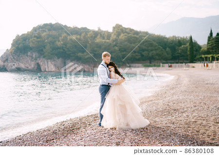 The bride and groom stand on the pebbled seashore and hug, the groom hugs the bride's shoulders The bride and groom stand on the pebbled seashore and hug, the groom hugs the bride's shoulders 86380108