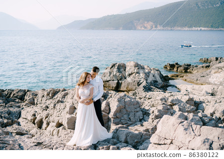 Groom hugs bride while standing on the stones against the background of the sea and mountains Groom hugs bride while standing on the stones against the background of the sea and mountains 86380122