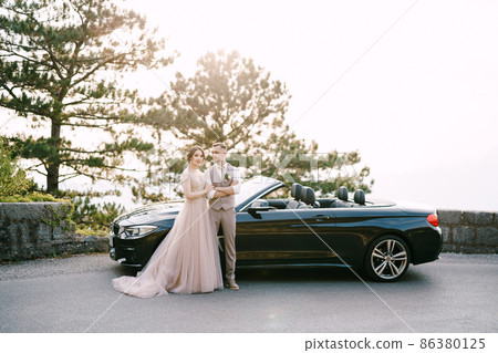 Bride and groom stand near the convertible on the road Bride and groom stand near the convertible on the road 86380125