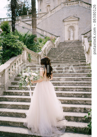 The bride stands on the stairs of the ancient church in Prcanj with a bridal bouquet in her hands, back view The bride stands on the stairs of the ancient church in Prcanj with a bridal bouquet in her hands, back view 86380126