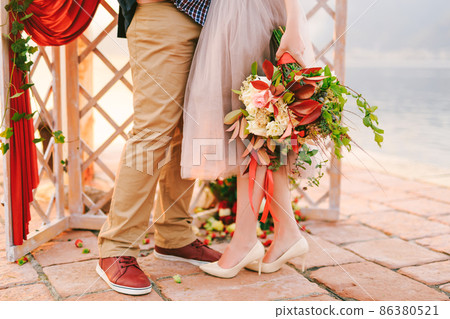 Groom and bride with a bouquet of flowers stand on a stone tile against the background of a carved screen decorated with a red curtain and greenery. Close-up 86380521