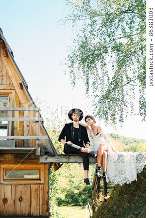 Bride and groom sit on a bridge near a house Bride and groom sit on a bridge near a house 86381003
