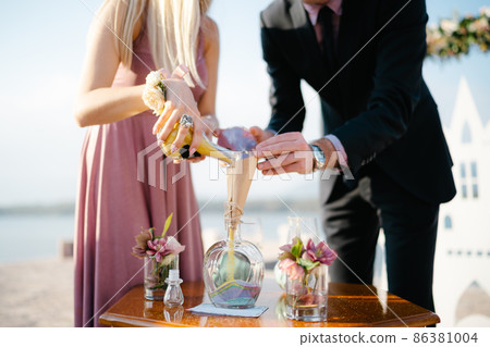 Groom and bride pour multi-colored sand in layers into a glass jar Groom and bride pour multi-colored sand in layers into a glass jar 86381004