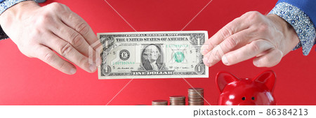 Male hands hold one dollar against the background of stacks of coins with red pig piggy bank Male hands hold one dollar against the background of stacks of coins with red pig piggy bank 86384213