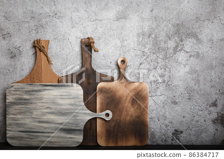 Samples of four different handmade rustic wooden cutting boards on a kitchen countertop against a concrete wall. Manufacture of household goods and accessories made of wood 86384717