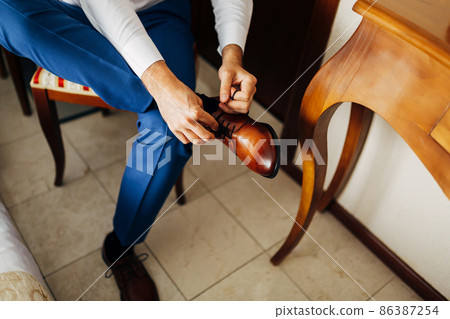 Man in a white shirt and blue trousers ties his boots, sitting on a chair. Close-up 86387254