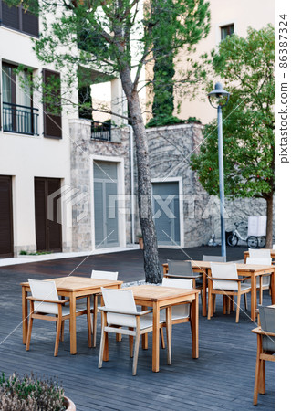Wooden tables and chairs on a wood terrace in a street cafe near a stone house 86387324