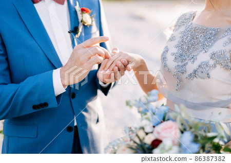 Groom in a blue suit puts a wedding ring on the bride's finger with a bouquet of flowers. Close-up 86387382