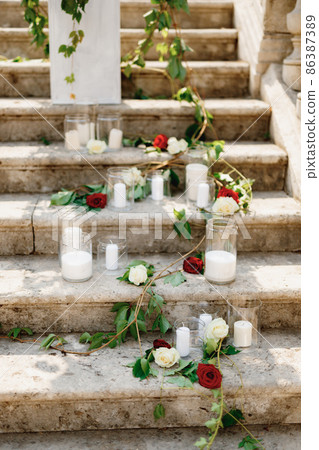 White candles in glass jars decorated with roses and green branches on stone steps White candles in glass jars decorated with roses and green branches on stone steps 86387389
