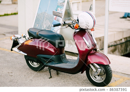 Burgundy motor scooter with a white helmet stands on the street Burgundy motor scooter with a white helmet stands on the street 86387391