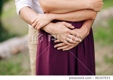 Hands of the groom in a white shirt hug from behind bride in a burgundy dress. Close-up Hands of the groom in a white shirt hug from behind bride in a burgundy dress. Close-up 86387633