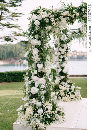 Wedding arches decorated with white flowers and green branches stand on a wooden platform Wedding arches decorated with white flowers and green branches stand on a wooden platform 86387698