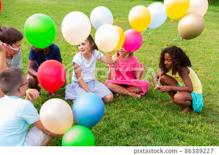 Cheerful preteen children sitting on lawn with balloons and talking 86389227