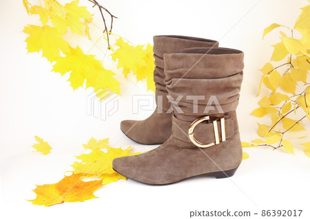 Beige brown suede short wide ankle buckle boots without heels with golden metal buckle and yellow autumn maple leaves on white background. Studio shot photo, front view, closeup. Selective focus. Beige brown suede short wide ankle buckle boots without heels with golden metal buckle and yellow autumn maple leaves on white background. Studio shot photo, front view, closeup. Selective focus. 86392017