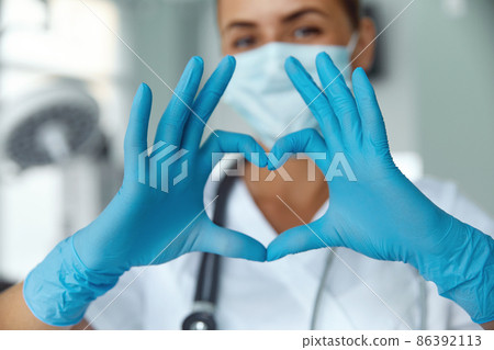 Woman with a medical mask and hands in latex glove shows the symbol of the heart. Doctor for the heart. Love our medical professionals. 86392113