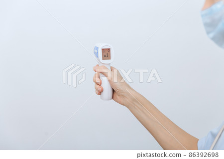 Temperature measurement gun in doctor hands. Close-up shot of doctor wearing protective surgical mask ready to use infrared isometric thermometer gun to check body temperature for virus symptoms. 86392698