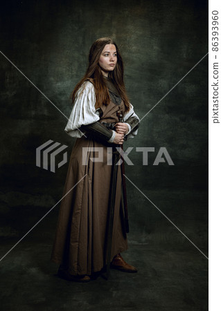 Full-length portrait of seriuos beautiful woman in image of medieval warrior or knight with dirty wounded face with big sword isolated over dark vintage background. 86393960