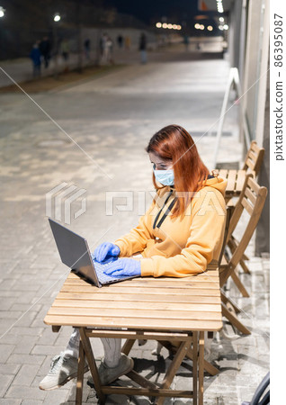 A woman in a medical mask and gloves remotely works late at night outdoors. Ocher-colored hoodie girl with laptop on the street during quarantine. Coronavirus. 86395087