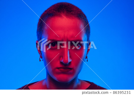 Close-up angry girl, student with short hair looking at camera isolated on dark blue background in neon light. Concept of emotions, gender x 86395231