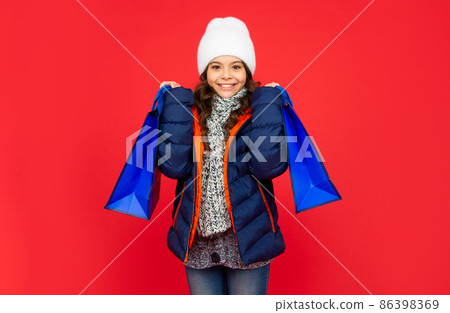happy kid in puffer jacket and hat. teen girl after shopping on red background. happy kid in puffer jacket and hat. teen girl after shopping on red background. 86398369