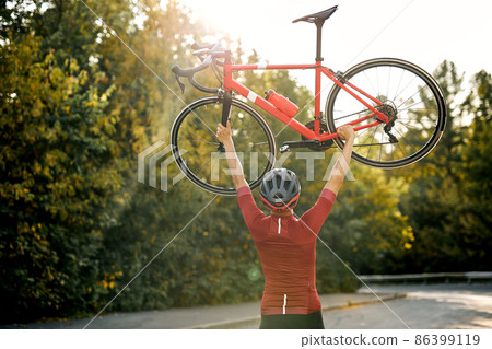 View from back on man in sportive clothes raising bike up after successful competition View from back on man in sportive clothes raising bike up after successful competition 86399119