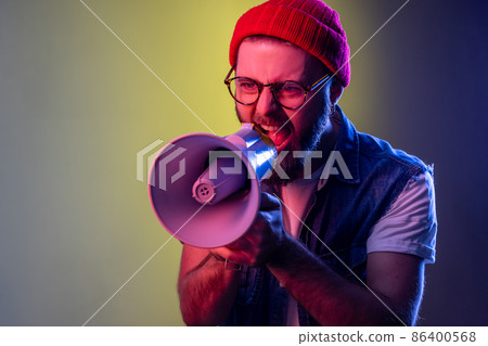Excited hipster guy screaming loud using megaphone, making announcement, presentation, protesting, wearing beanie hat and denim vest. Indoor studio shot isolated on colorful neon light background. 86400568