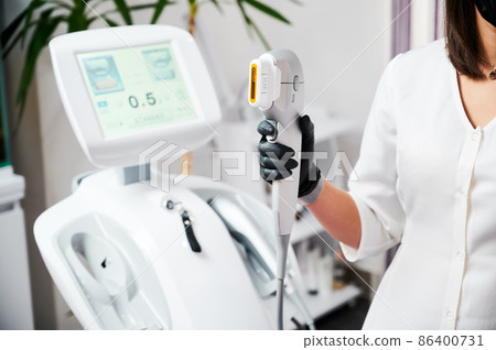 Close up of beautician hand in black glove holding ultrasound device for face lifting and skin tightening procedure. Young woman cosmetologist using modern cosmetology equipment in clinic. Close up of beautician hand in black glove holding ultrasound device for face lifting and skin tightening procedure. Young woman cosmetologist using modern cosmetology equipment in clinic. 86400731
