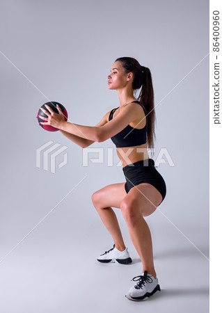 Side view portrait of Slim caucasian cross fit woman with fitness ball doing squats, isolated Side view portrait of Slim caucasian cross fit woman with fitness ball doing squats, isolated 86400960