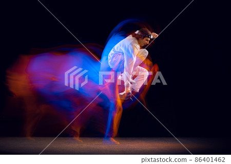 Flexible man in sports white suit dancing hip-hop isolated on dark background in mixed neon light. Youth culture, hip-hop, movement, style and fashion, action. Flexible man in sports white suit dancing hip-hop isolated on dark background in mixed neon light. Youth culture, hip-hop, movement, style and fashion, action. 86401462