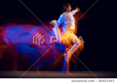 Youth dances. Dynamic portrait of hip-hop dancer in action, motion isolated on dark background in mixed neon light. Youth culture, hip-hop, style and fashion 86401463
