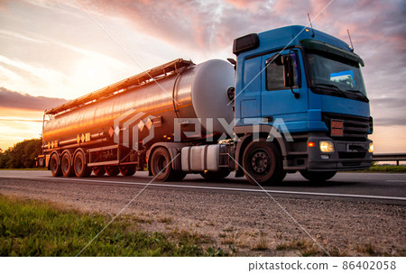 A blue modern truck with a semi-trailer tanker transports dangerous goods against the backdrop of the evening sunset. The concept of transportation of liquid cargo, hazard class ADR. Industry A blue modern truck with a semi-trailer tanker transports dangerous goods against the backdrop of the evening sunset. The concept of transportation of liquid cargo, hazard class ADR. Industry 86402058