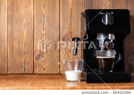 A cup of black coffee and a cup of milk stand near a coffee maker with a manual cappuccino maker. Wood background. Copy space for text, natural 86402086