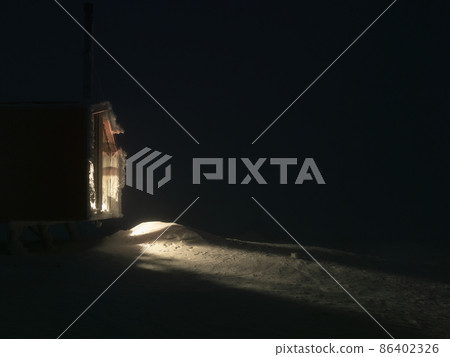 Side light from the night window of the cottage on a winter meadow with snowdrifts. 86402326