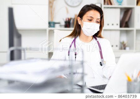 Woman doctor in protective mask sitting at workplace with computer in office 86403739