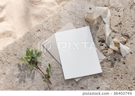 Summer wedding stationery mock-up. Blank greeting card, invitation. Envelope and silk ribbon in sunlight. Green lentisk branch. Beige concrete background in sunlight. Blurred sandy beach. Top view. 86403749