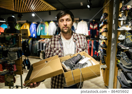 Caucasian male salesman consultant in outdoor store presenting hiking boots to buyer. A salesperson at a sports store helps a shopper to choose hiking boots. Seller in sporting goods store 86405602