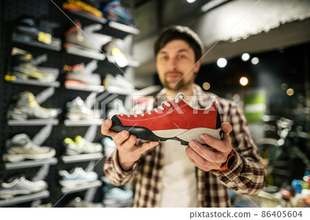 Salesman advises client on choice of hiking shoes in an outdoor store. Male tourist shop owner helping customer try on hiking boots. Young seller confidently presents mountain boot. sell, trade 86405604
