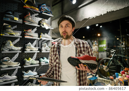 Salesman advises client on choice of hiking shoes in an outdoor store. Male tourist shop owner helping customer try on hiking boots. Young seller confidently presents mountain boot. sell, trade Salesman advises client on choice of hiking shoes in an outdoor store. Male tourist shop owner helping customer try on hiking boots. Young seller confidently presents mountain boot. sell, trade 86405617