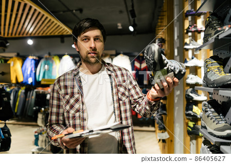 Caucasian male salesman consultant in outdoor store presenting hiking boots to buyer. A salesperson at a sports store helps a shopper to choose hiking boots. Seller in sporting goods store 86405627