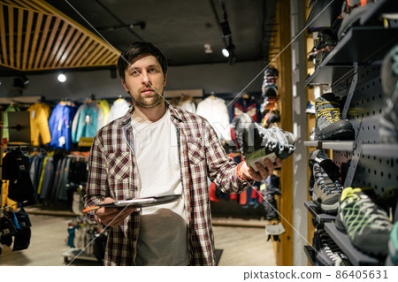 Store clerk helping customer try hiking boots at outdoors equipment shop. Salesman demonstrating trekking and hiking footwear for outdoor trails. Consultant help choose hiking boots in sports store 86405631