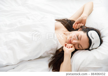 Happy awakening of a young woman from a dream. A woman in bed wearing a sleep mask stretches, looks at the camera and smiles. The concept of good morning. Happy awakening of a young woman from a dream. A woman in bed wearing a sleep mask stretches, looks at the camera and smiles. The concept of good morning. 86406432