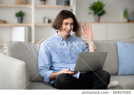Distant communication concept. Happy man video calling on laptop computer, waving hand to screen, sitting on sofa Distant communication concept. Happy man video calling on laptop computer, waving hand to screen, sitting on sofa 86407098