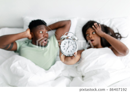 Shocked millennial black male and female with open mouths show alarm clock, lies on white bed in bedroom Shocked millennial black male and female with open mouths show alarm clock, lies on white bed in bedroom 86407192
