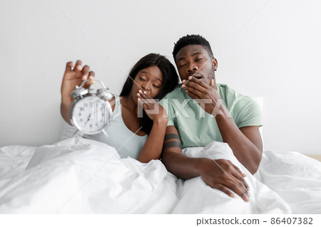 Tired sad sleepy millennial african american couple sit on white bed in bedroom, yawn and show alarm clock Tired sad sleepy millennial african american couple sit on white bed in bedroom, yawn and show alarm clock 86407382