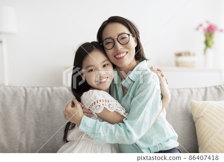 Happy little Asian girl and her loving grandmother hugging and looking at camera on couch at home 86407418